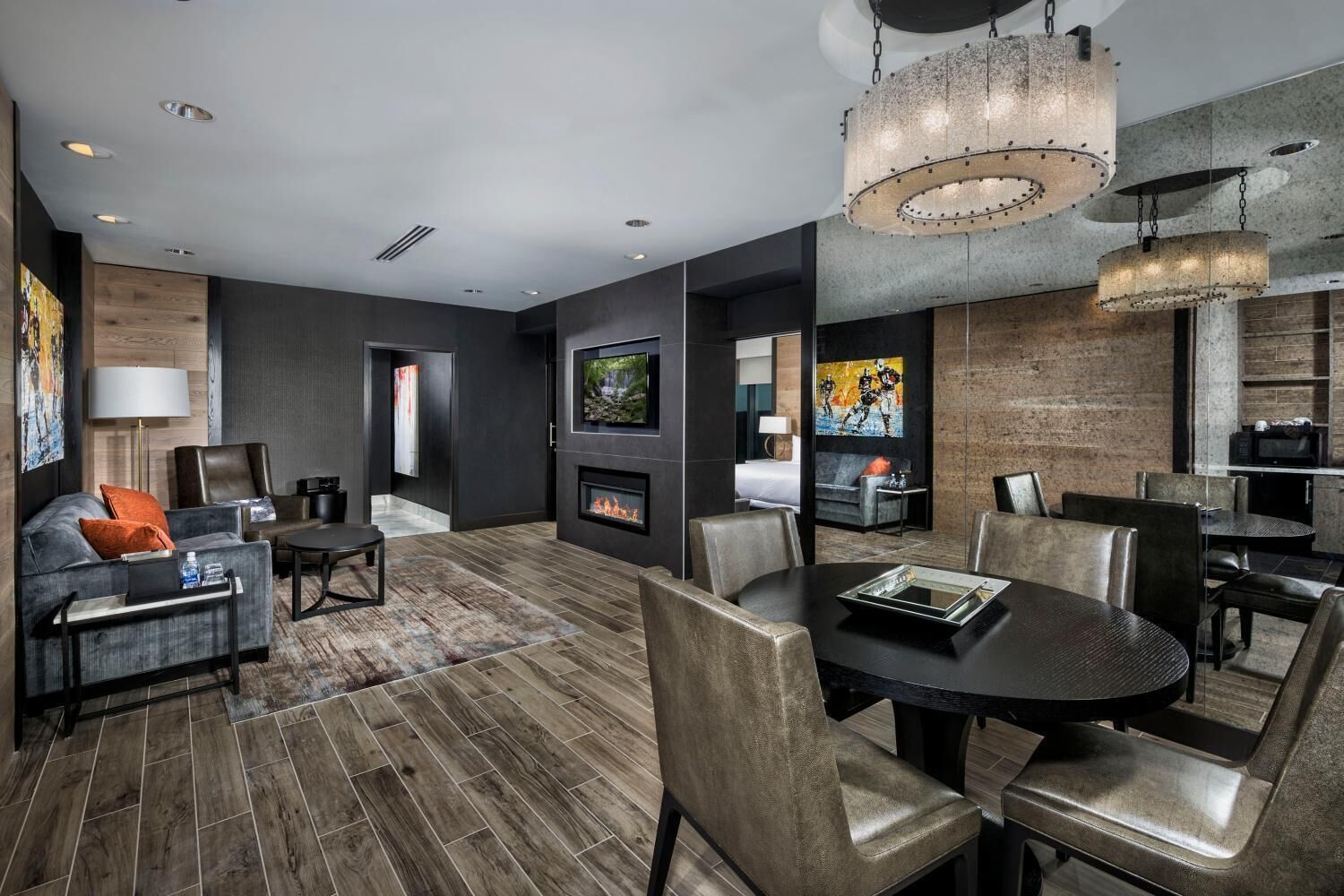 A modern hotel room with a dark wood floor, a fireplace, a dining area with a round table, and a large chandelier.