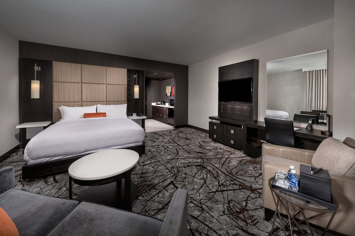 A modern hotel room with a large bed, a small round table, a sofa, a TV, and a minibar.