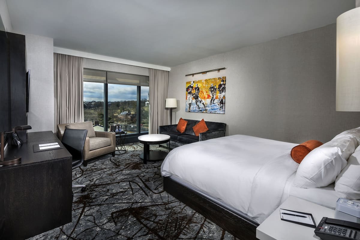 A hotel room with a large bed, a desk, a sofa, and a small table. There is a painting on the wall and a window with curtains.