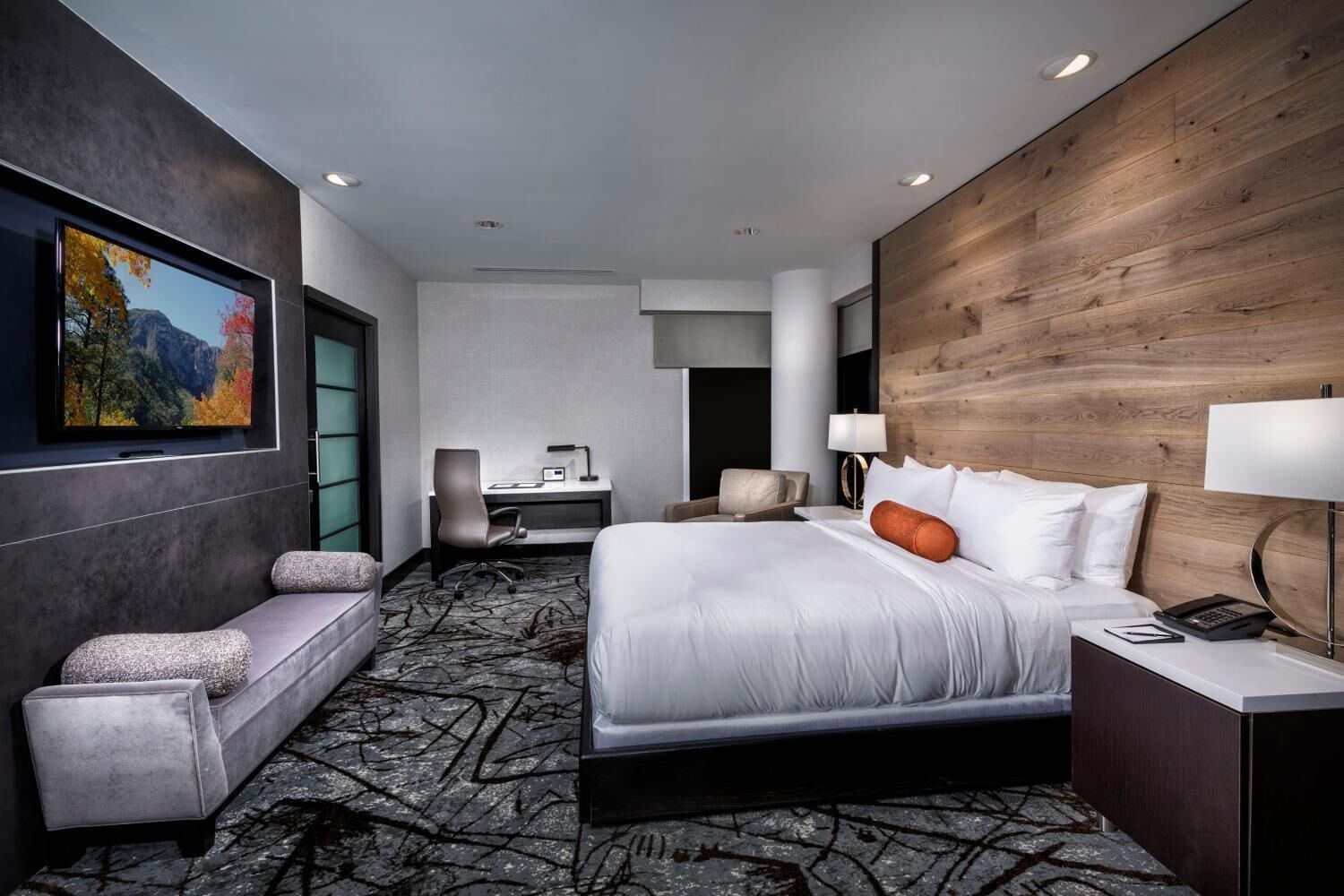 A modern hotel room with a large bed, a desk with a chair, a sofa, and a TV on the wall.