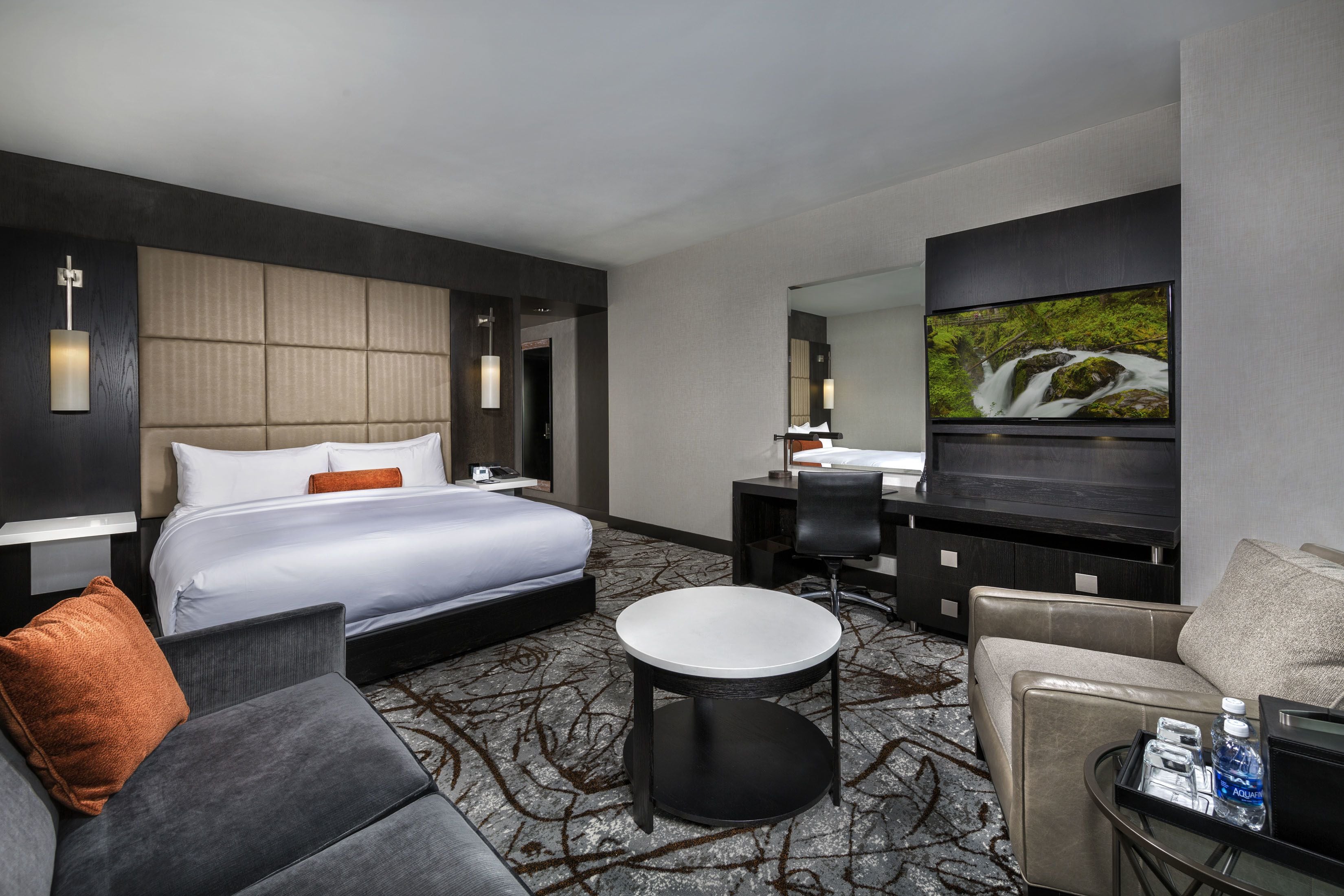 A modern hotel room with a large bed, a sitting area with a sofa and armchair, a small round table, a desk with a chair, and a flat-screen TV mounted on the wall.