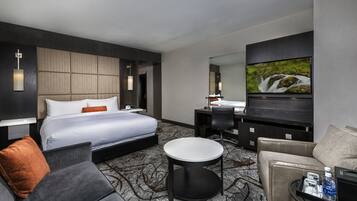 A modern hotel room with a large bed, a sitting area with a sofa and armchair, a small round table, a desk with a chair, and a flat-screen TV mounted on the wall.