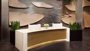 A reception desk with a marble top and a wooden base, flanked by two black planters with green plants.