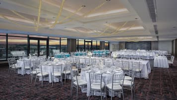 Un salón de banquetes con mesas redondas, manteles blancos y sillas de plata, con vistas a la ciudad a través de grandes ventanales.
