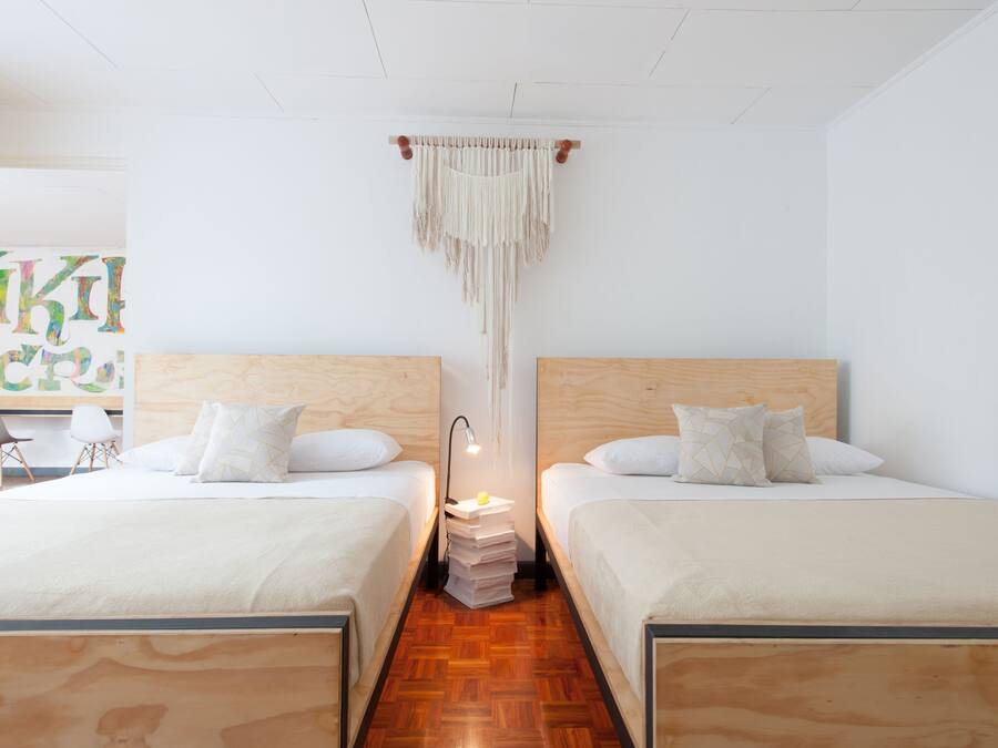 A hotel room with two beds, wooden headboards, white bedding, a small bedside table with a lamp, and a macrame wall hanging.