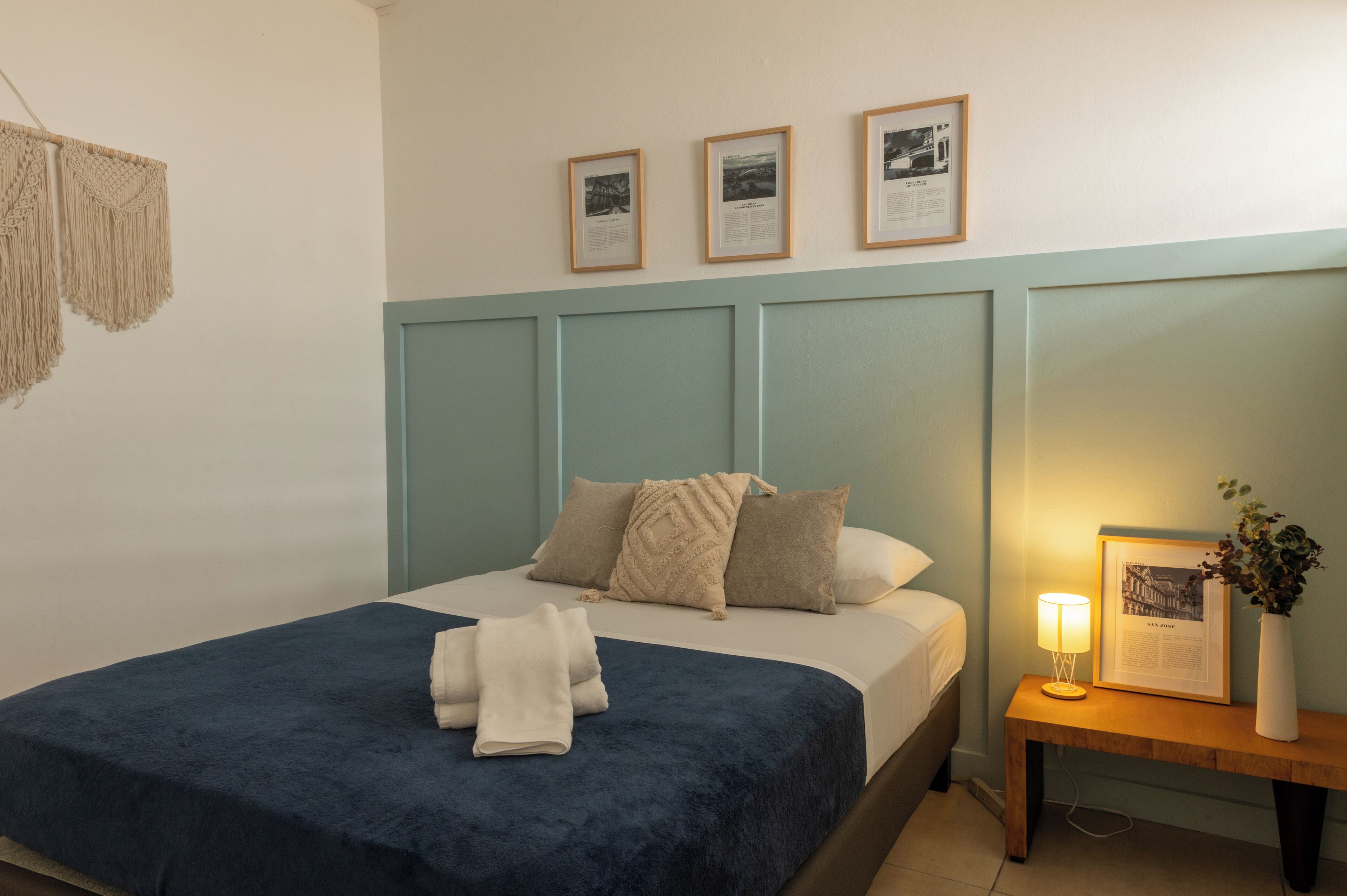 A neatly made bed with a blue bedspread and white towels, a bedside table with a lamp and framed pictures, and macramé wall hangings.