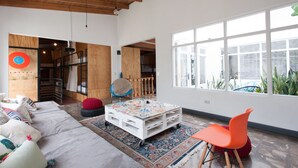 A spacious living room with a large window, a white sofa, colorful cushions, a white coffee table, and a red chair. - Capital Hostel de Ciudad (San José)