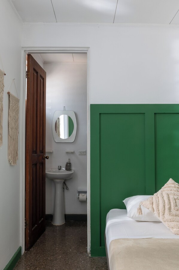 Una habitación con cabecera verde, cama blanca, puerta de madera, lavamanos blanco y un espejo.