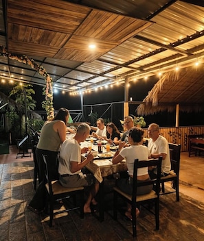 Un groupe de personnes est assis à une table en plein air, sous un toit de chaume, et des guirlandes lumineuses illuminent les lieux.
