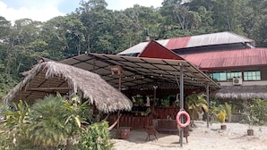 A rustic building with a thatched roof and a red metal roof, surrounded by lush greenery and a sandy ground. - Grand Selva Lodge & Tours (Ahuano)