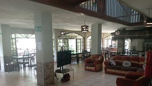 A spacious lobby with a high ceiling, tiled floor, and a seating area with sofas and armchairs. There is a television on a stand, a fire extinguisher, and a staircase leading to an upper level. - Grand Selva Lodge & Tours (Ahuano)