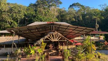 A covered outdoor area with a thatched roof, surrounded by lush greenery and trees.