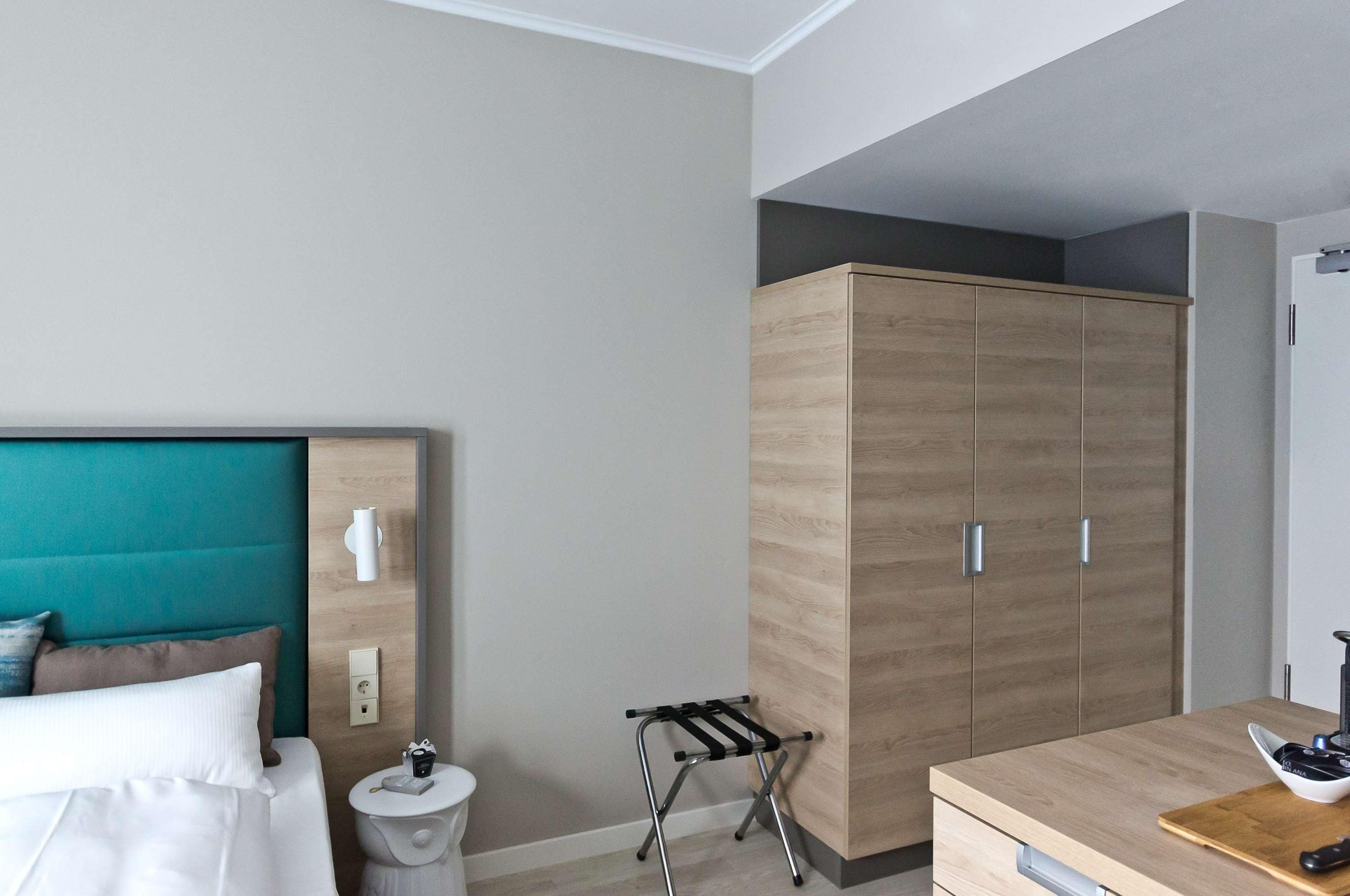 Une chambre d’hôtel comprenant un lit, une armoire en bois, un petit tabouret, une table de chevet et un bureau en bois avec une chaise.