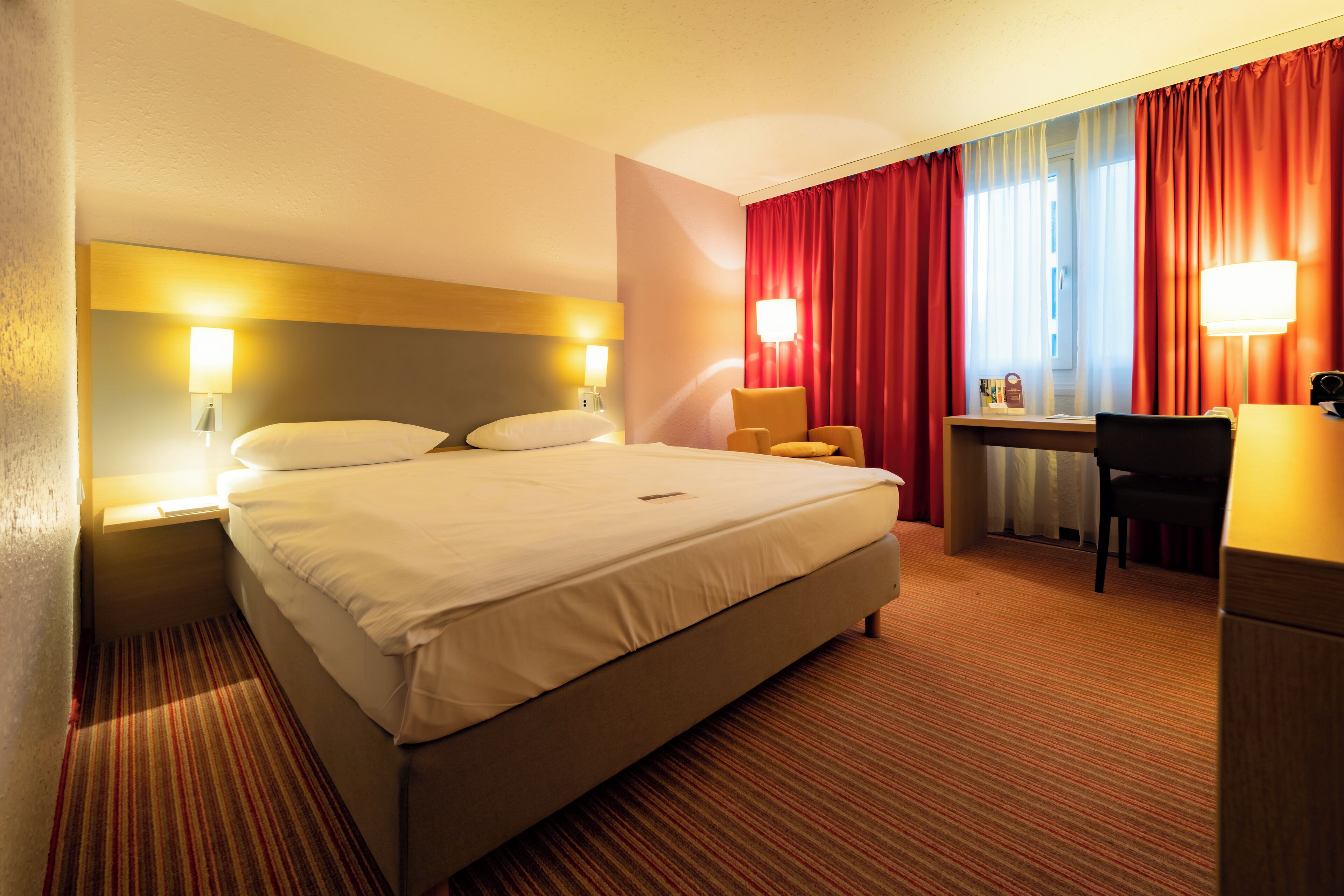 A hotel room with a bed, a desk, a chair, and a lamp.