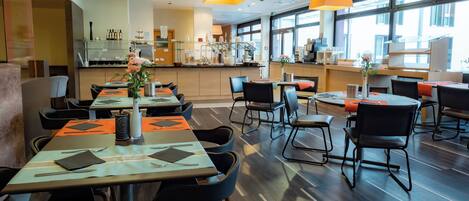 A modern restaurant with multiple dining tables and chairs, large windows, and hanging pendant lights.