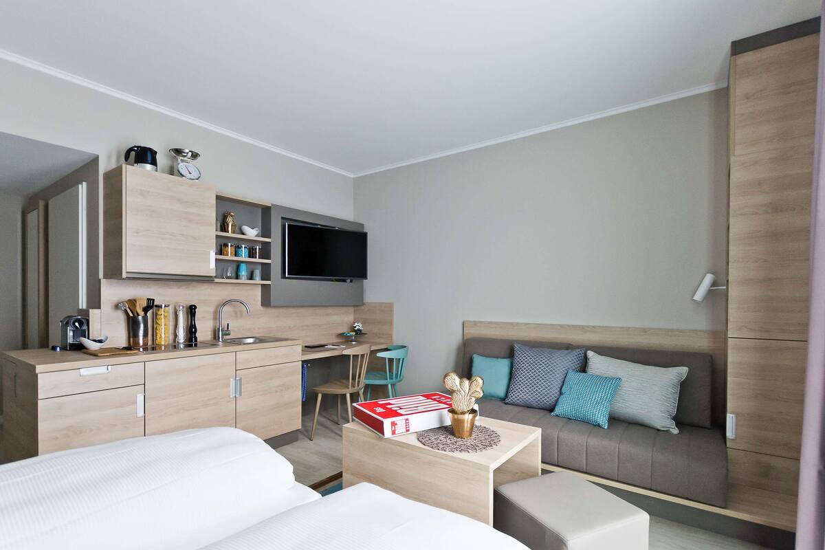 A modern hotel room with a kitchenette, a sofa, a TV, and a dining area.