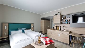 A modern hotel room with a bed, bedside table, and a kitchenette area including a sink and a TV.