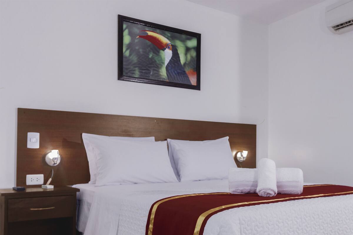 Una cama bien hecha con sábanas blancas y detalles en rojo y dorado, una mesita de noche con una lámpara y un cuadro con un tucán en la pared.