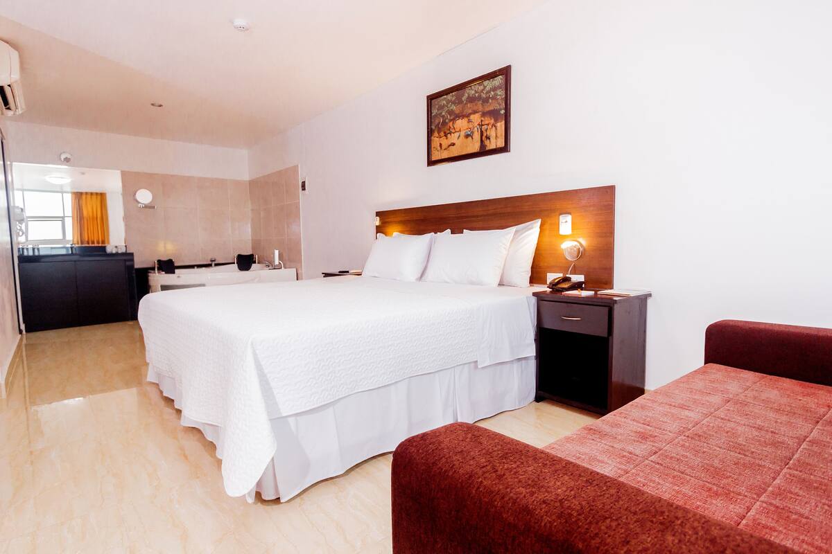Una habitación de hotel con una cama grande, cabecera de madera, mesita de noche, una silla roja y una pequeña mesa.