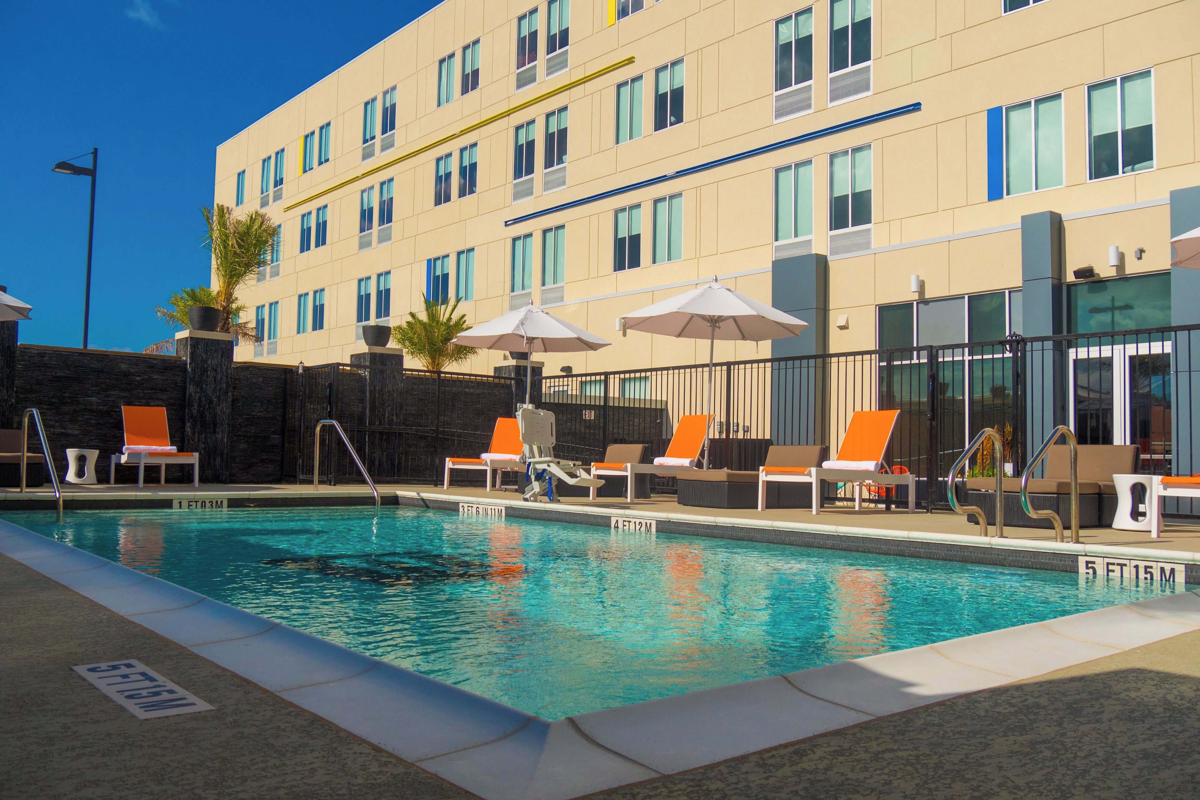 A hotel pool area with lounge chairs and umbrellas, a clear sky, and a building with multiple windows.