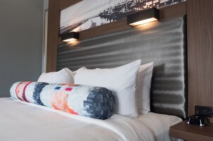 Een net opgemaakt bed met een kussen met patroon, een hoofdbord met ingebouwde verlichting en een digitaal klokje op het nachtkastje.