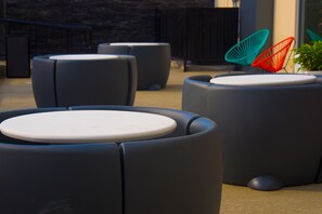 Outdoor seating with round tables and chairs, a potted plant, and a stone wall in the background.