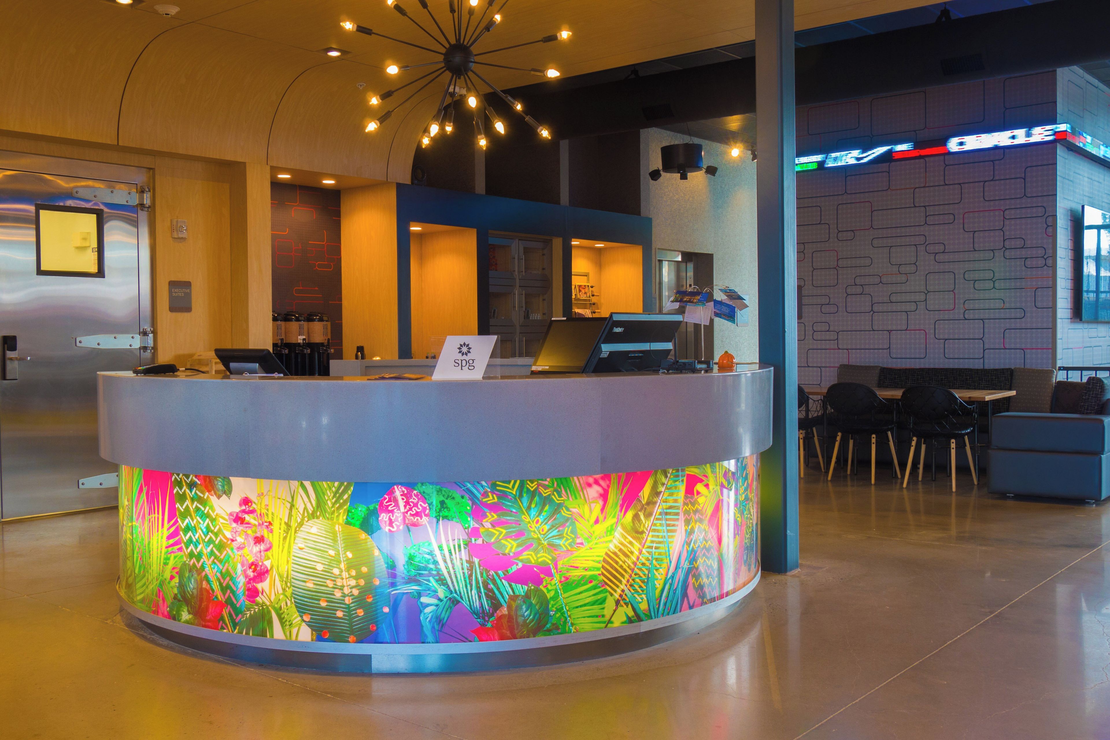 A reception desk with a colorful tropical-themed design, a modern chandelier, and a seating area with chairs and tables.