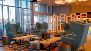 A modern lounge area with comfortable seating, a patterned rug, and a view of the outside through large windows. - Aloft by Marriott Corpus Christi (Corpus Christi)