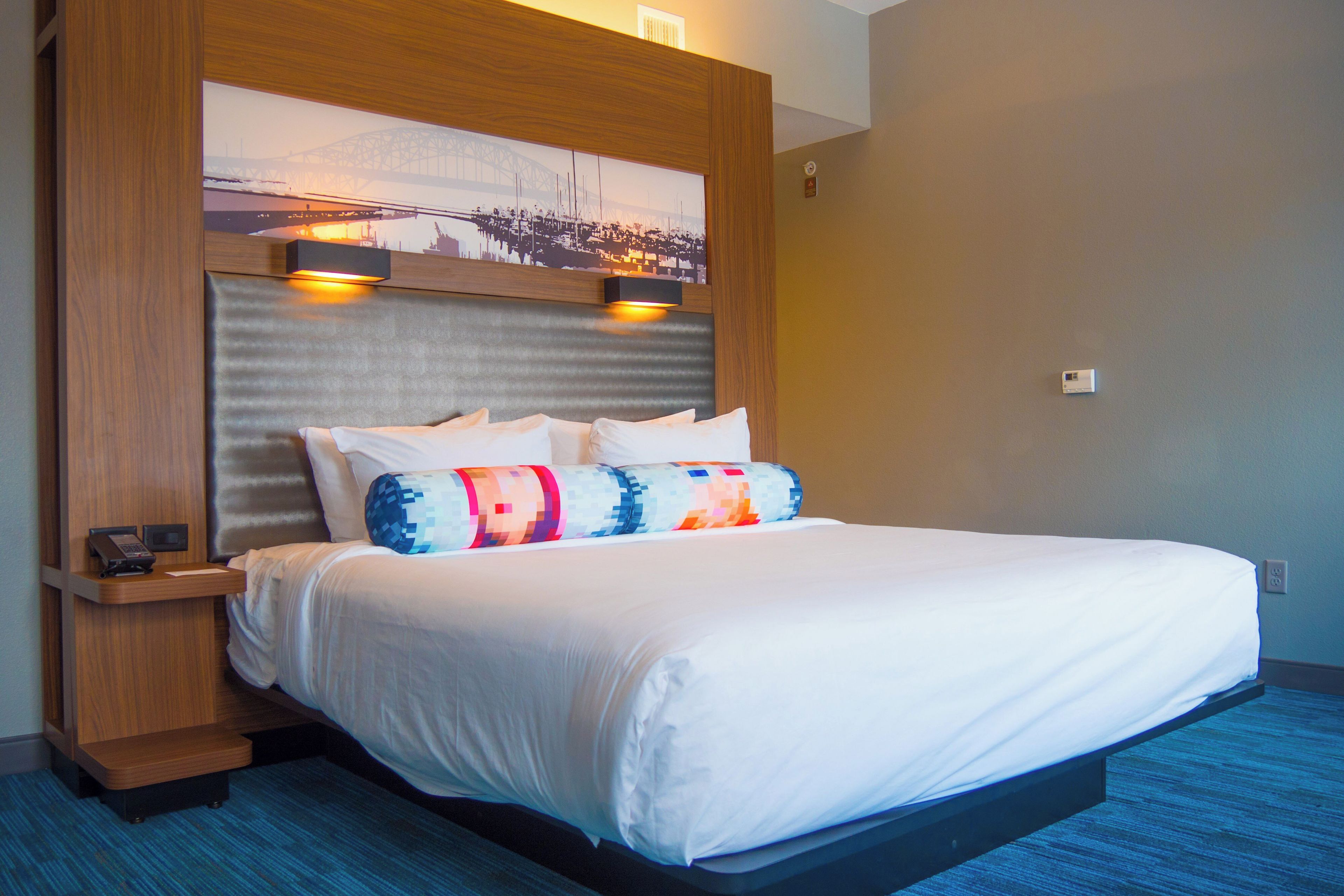 A hotel room with a large bed, headboard with integrated lighting, a nightstand with a phone, and a wall-mounted artwork.