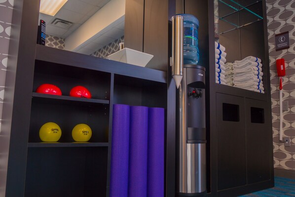 A hotel room with a water dispenser, a shelf with sports balls, a folded purple mat, and a stack of towels.
