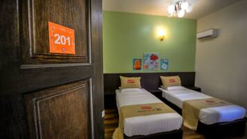 Une chambre d’hôtel avec deux lits simples, une porte en bois portant un numéro, un mur vert et un luminaire au plafond.