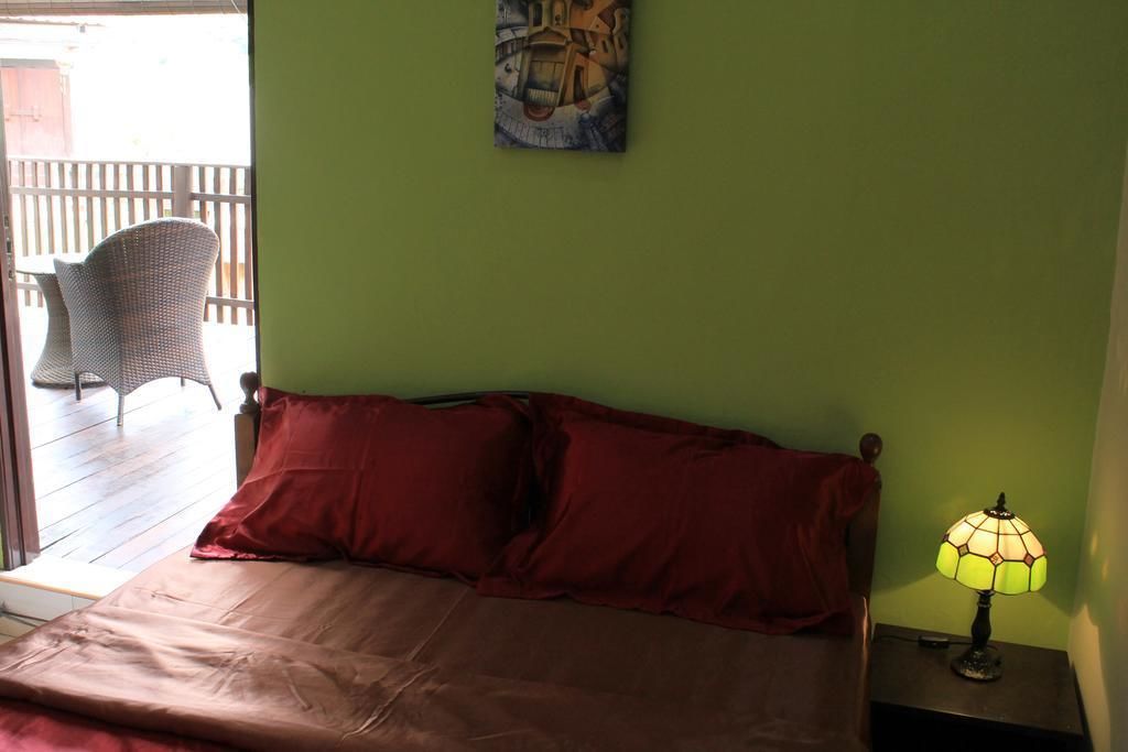 A room with a bed, a nightstand with a lamp, a painting on the wall, and a view of an outdoor patio with chairs.