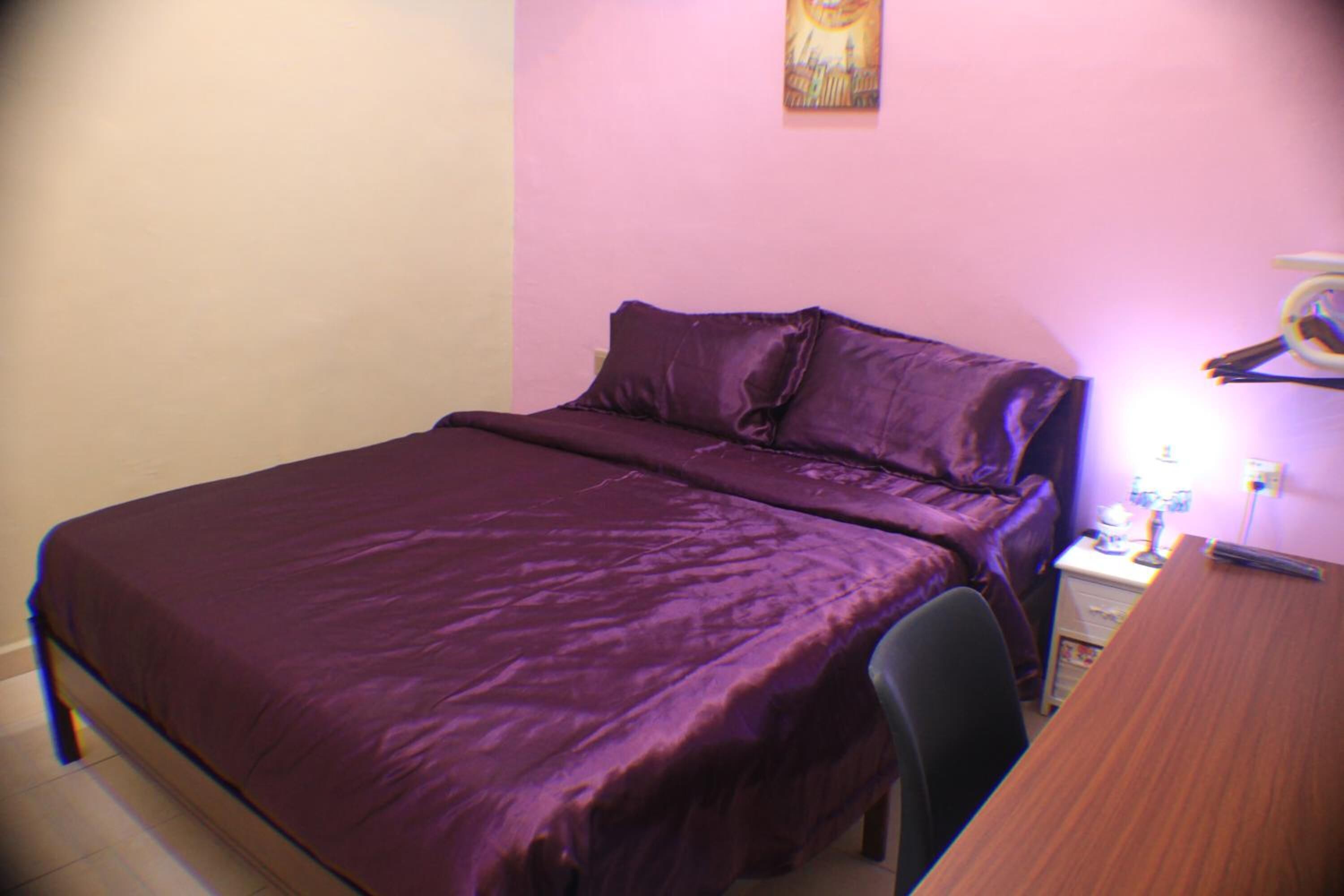 Una cama pequeña y bien hecha con cubrecama y almohadas moradas, una mesita de noche de madera con una lámpara, y una silla.