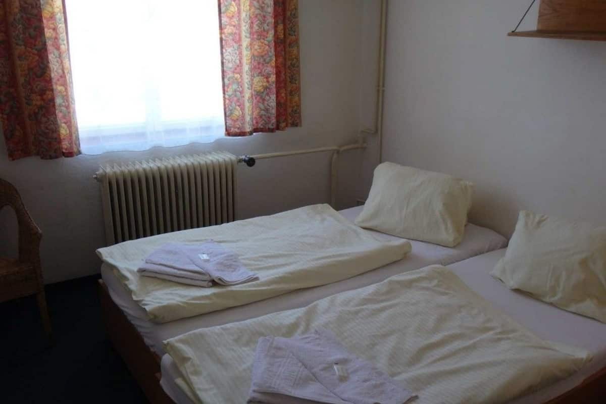 A room with two single beds, a radiator, a window with floral curtains, and a chair.