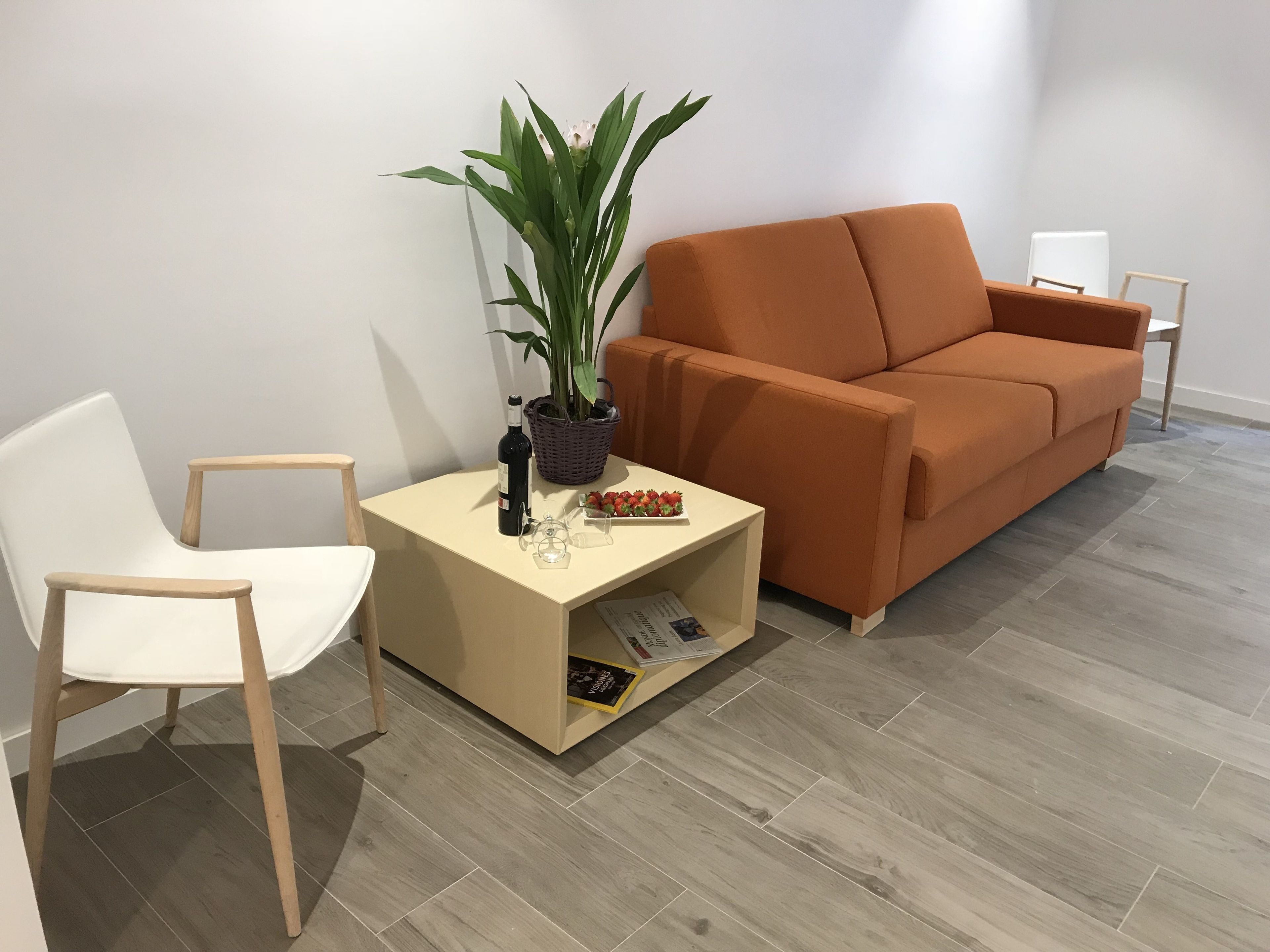 A modern living room with a white chair, an orange sofa, a small table with a plant, a wine bottle, and a basket.
