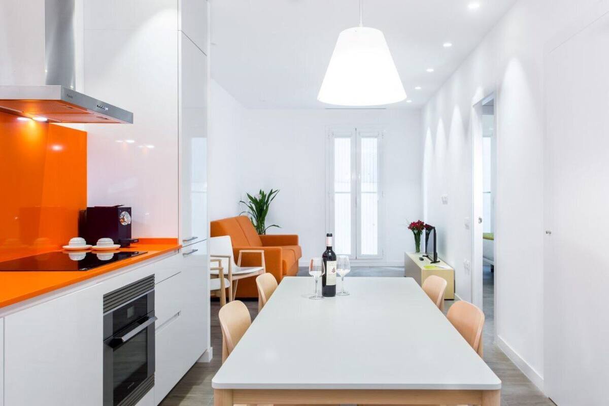 A modern kitchen with a white countertop and orange accents, a dining area with a white table and chairs, and a bright pendant light.