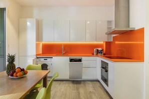 A modern kitchen with white cabinets, an orange countertop, and a wooden dining table with chairs. - Cosmo Apartaments Marina (Barcelona)