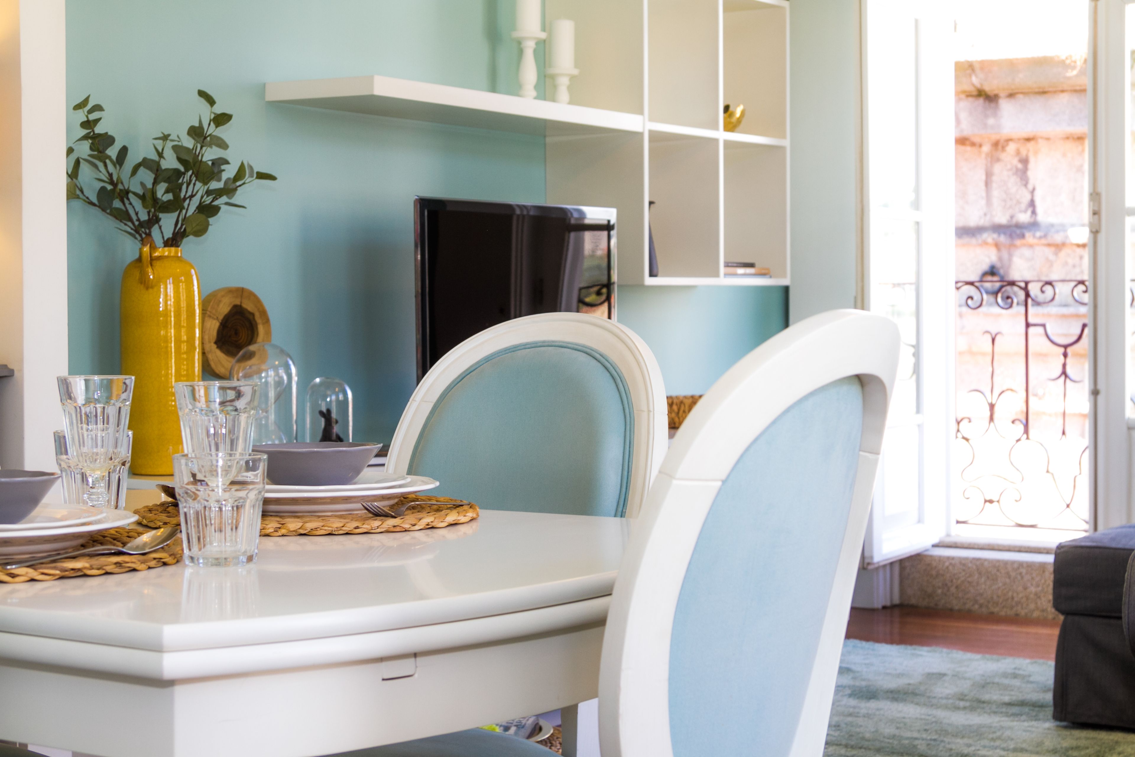 A dining area with a white table set for two, a yellow vase with green leaves, and a black refrigerator in the background.