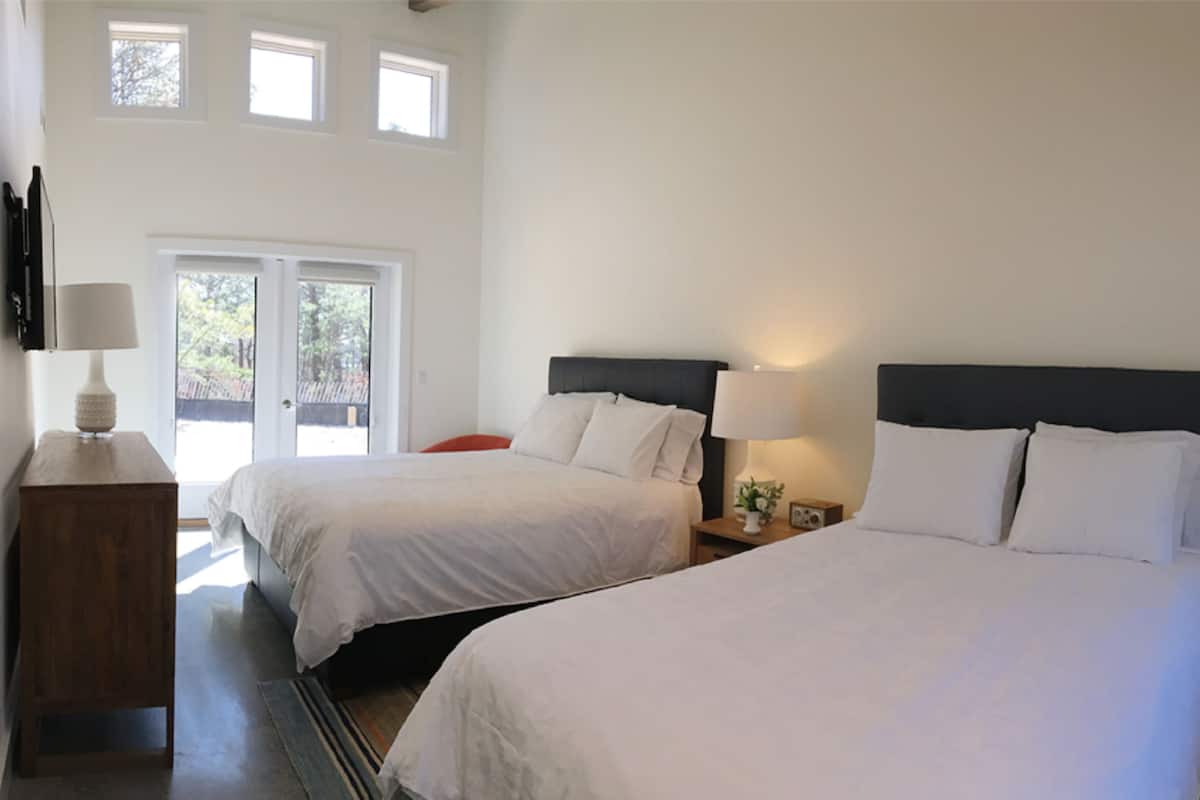 Habitación de hotel con dos camas, una mesita de noche de madera, una lámpara y vistas al exterior a través de una puerta de cristal.
