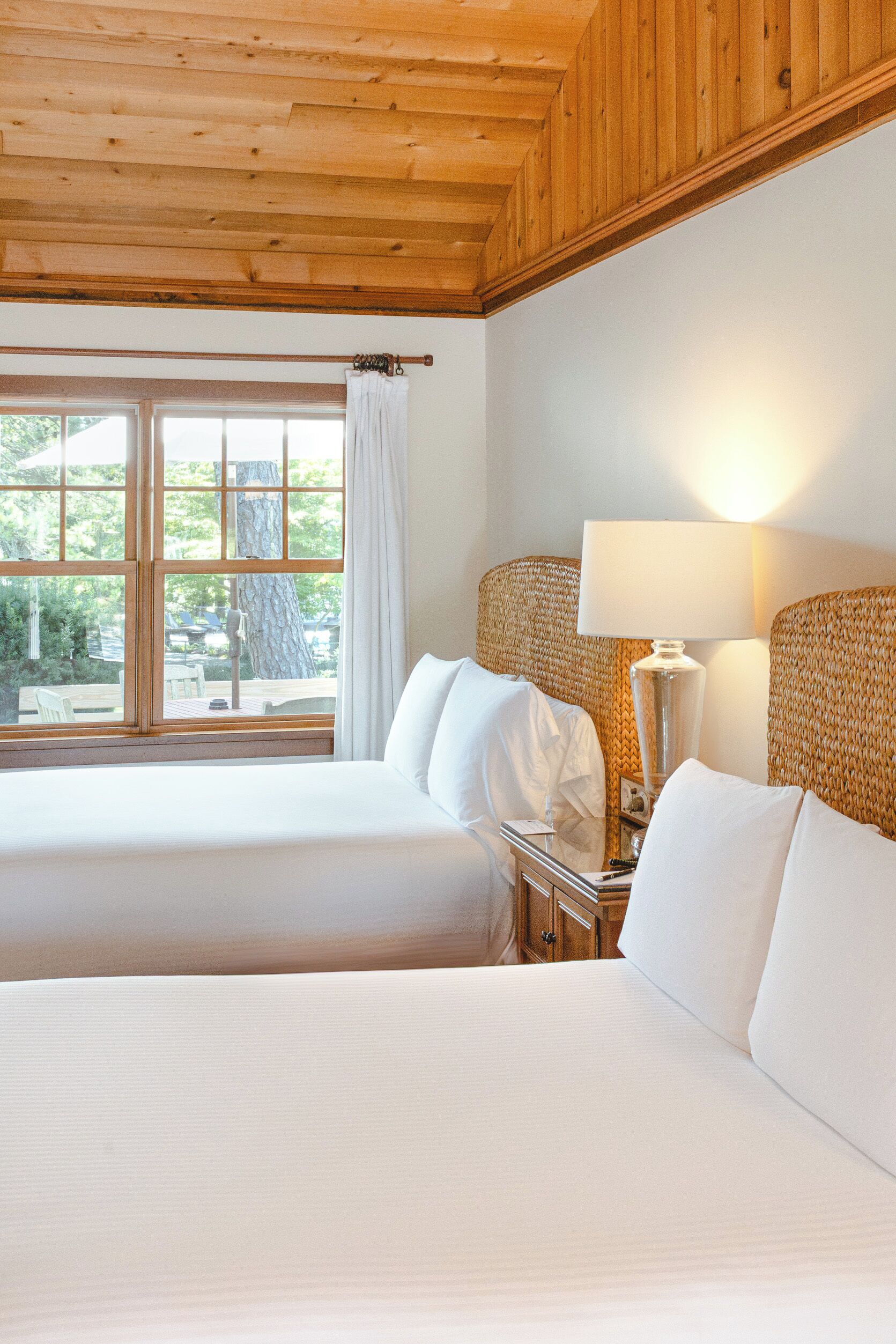 A hotel room with two beds, wooden ceiling, large windows with curtains, a bedside table with a lamp, and a view of the outdoors.