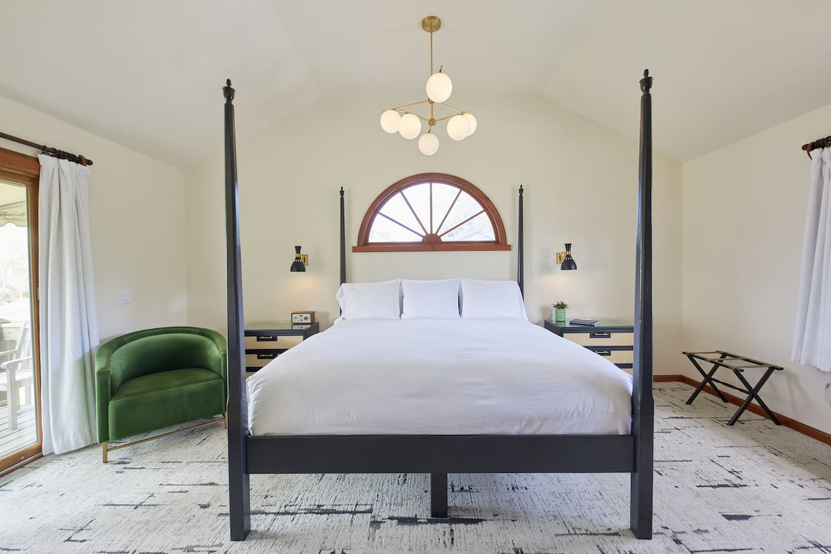 Una cama con dosel con una colcha blanca, un sillón verde, una ventana con marco de madera y un aplique colgante.