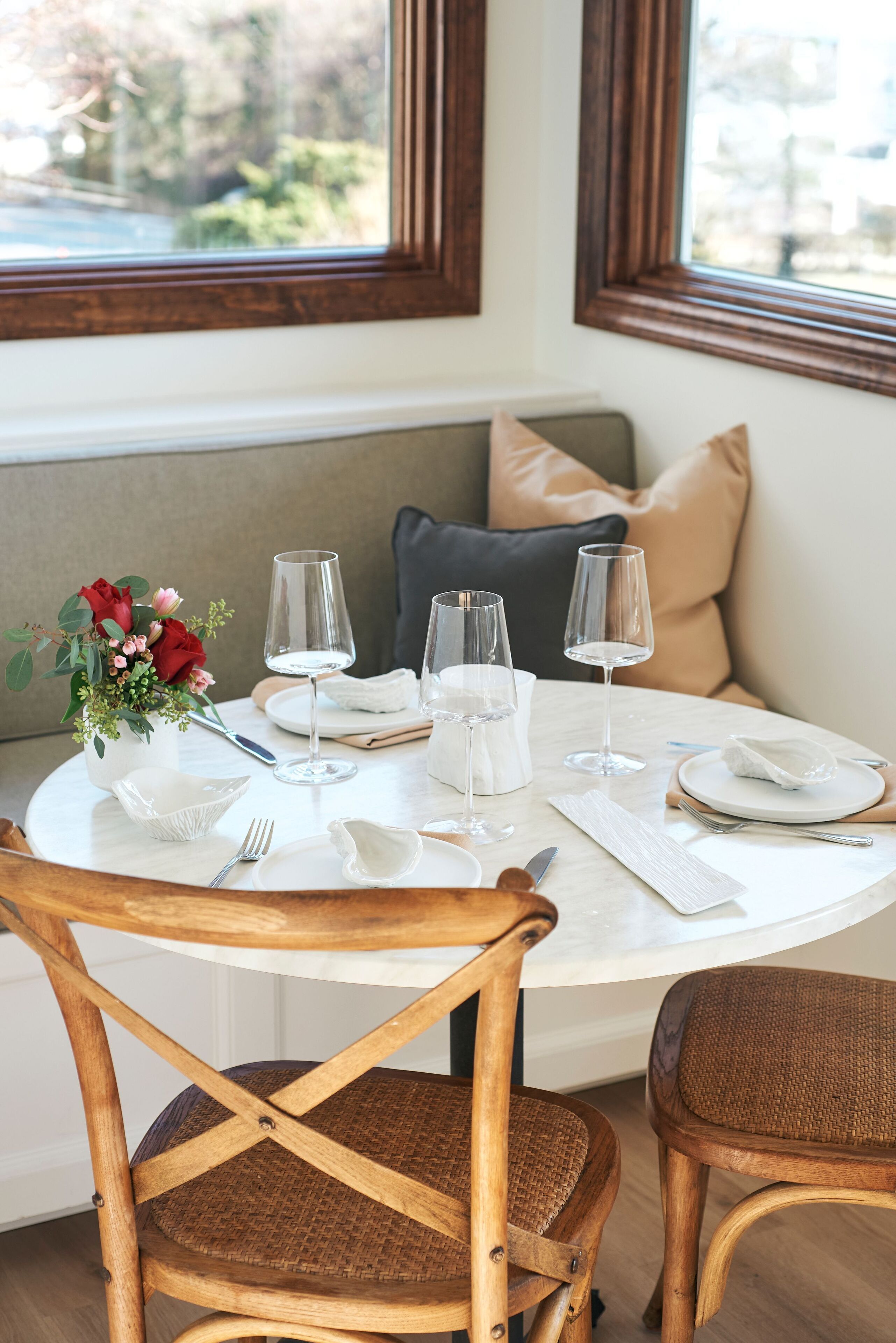 A round dining table set with white tableware, two wine glasses, and a vase with red roses.