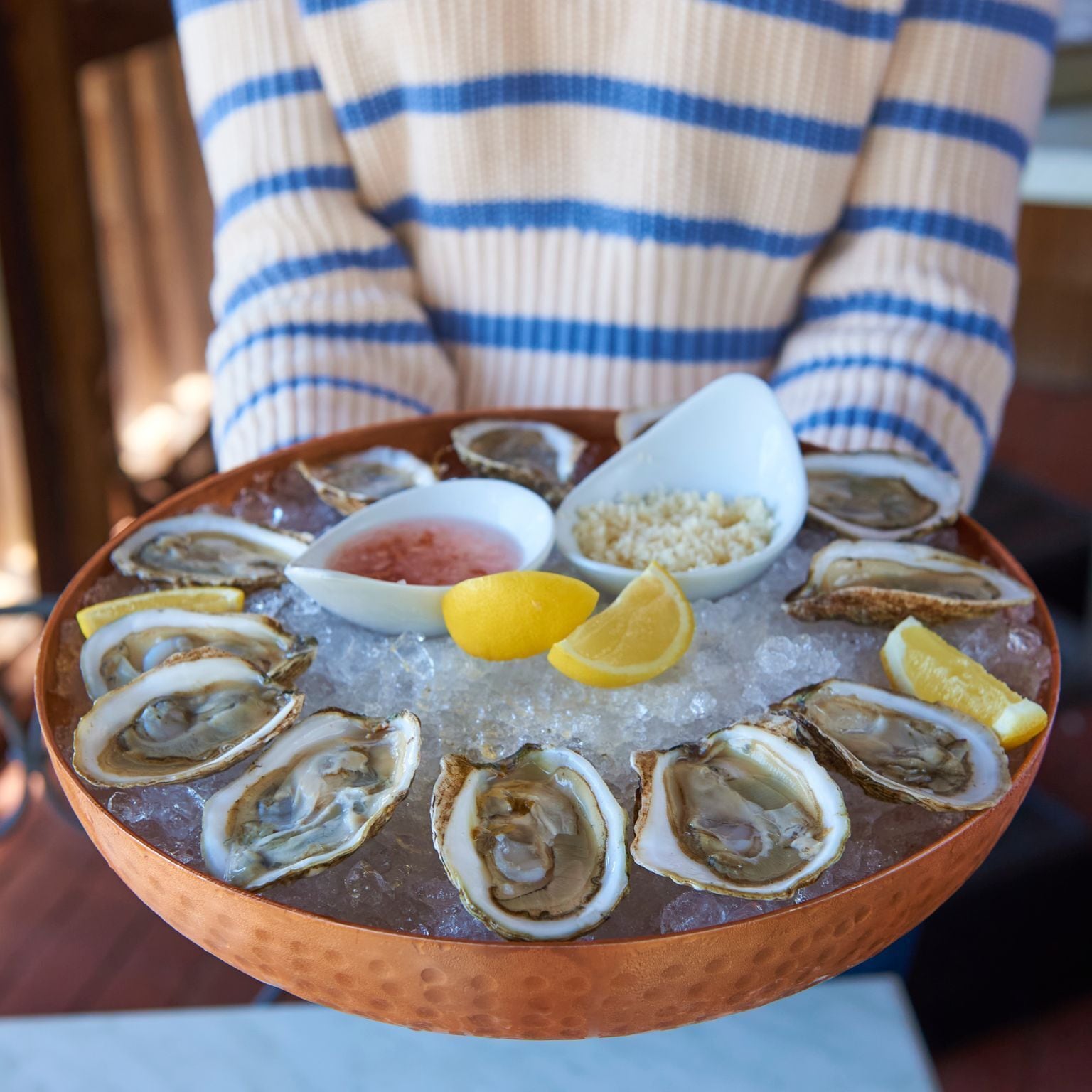 Een persoon houdt een koperen schaal vast met oesters op ijs, vergezeld van partjes citroen en een klein schaaltje saus.