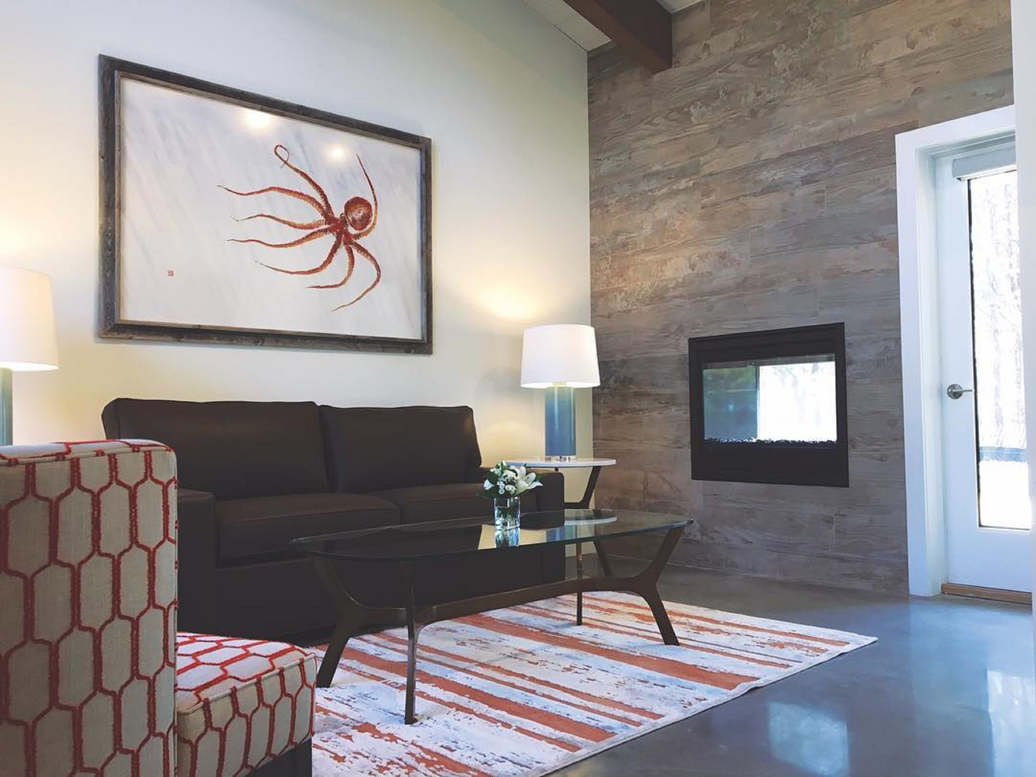 A modern living room with a dark brown sofa, a glass coffee table, and a patterned rug. A large abstract painting hangs on the wall, and a flat-screen TV is mounted above a wooden accent wall.