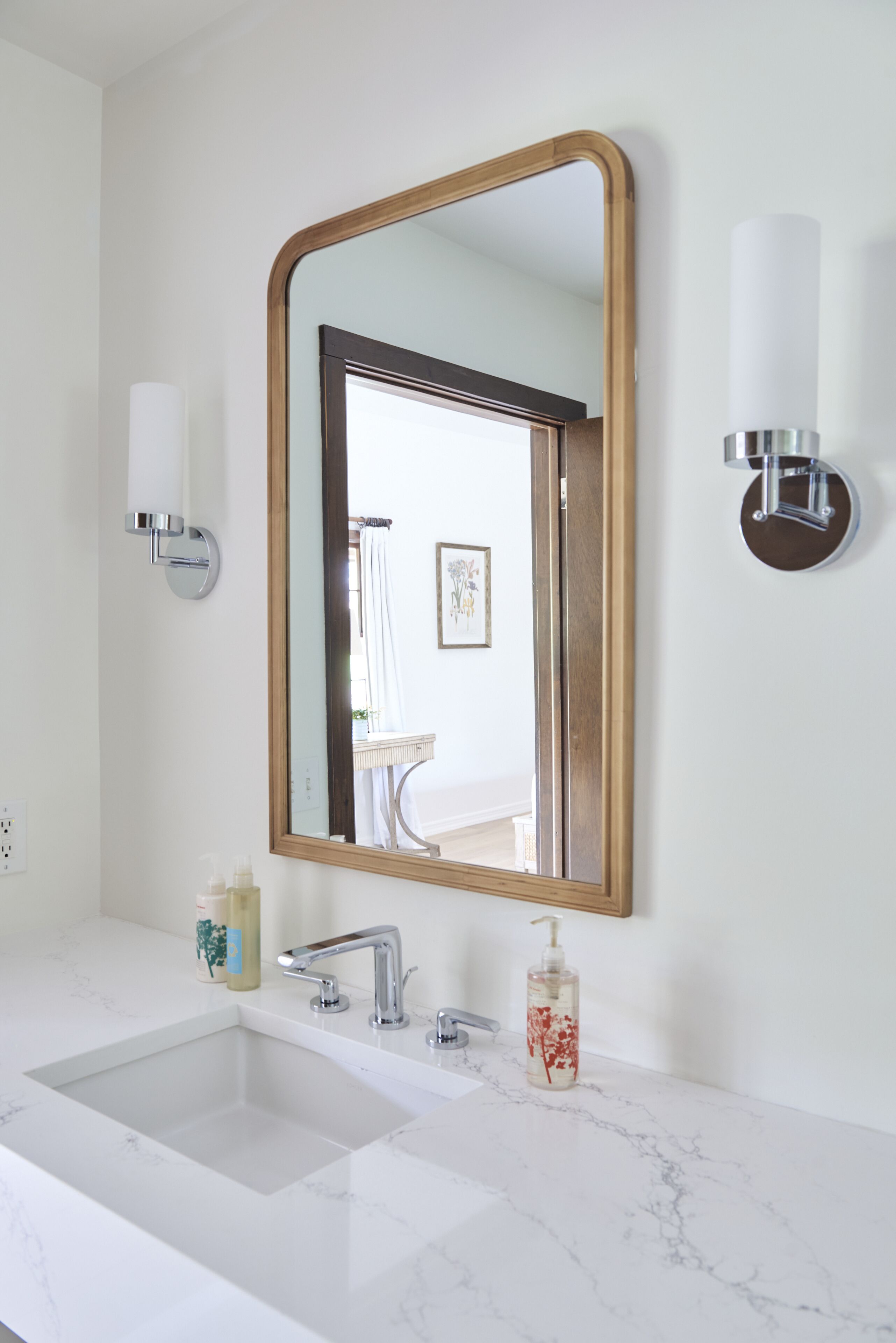 A bathroom with a white marble sink, a wooden-framed mirror, and a wall-mounted light fixture.