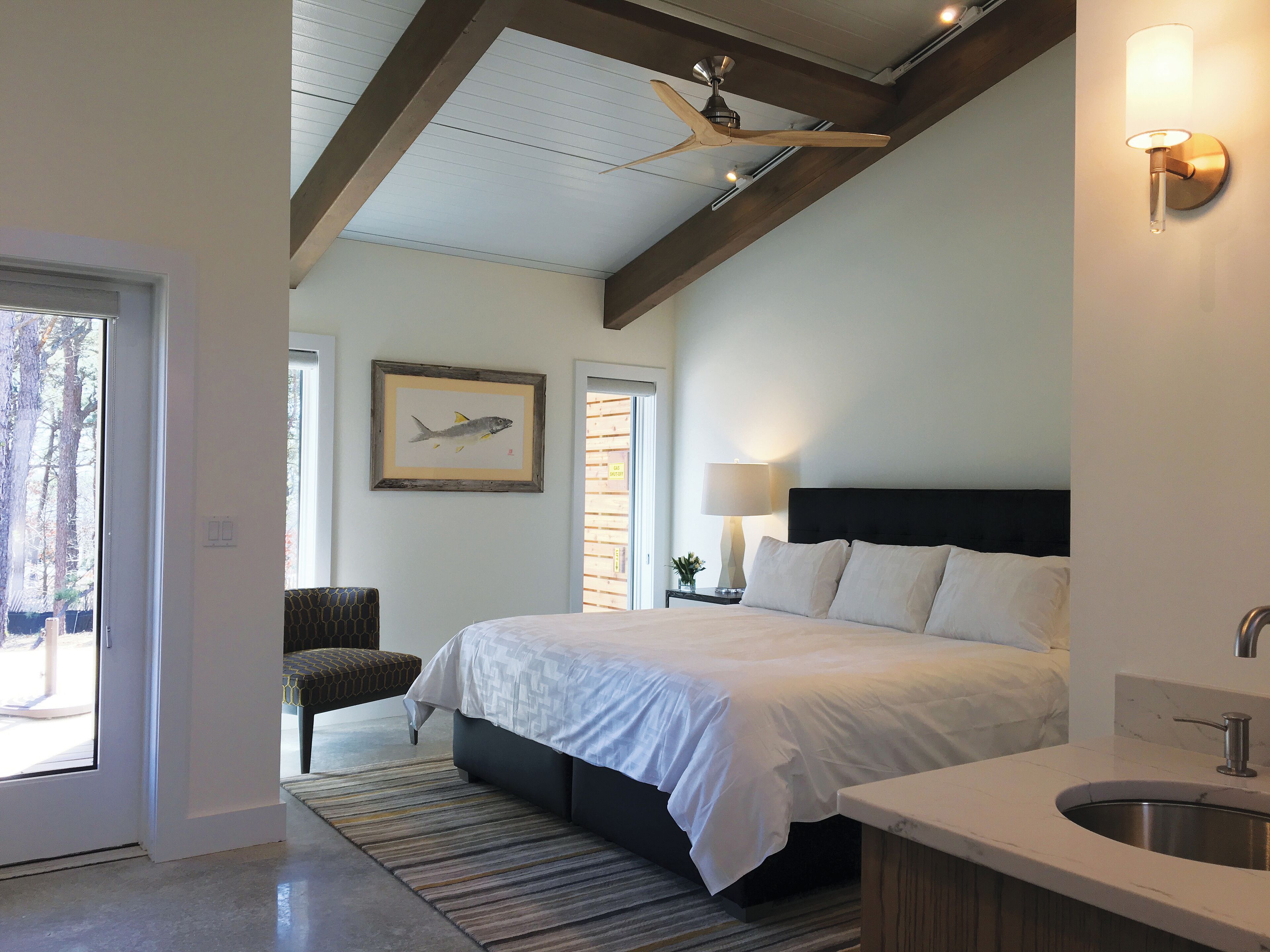 A bedroom with a bed, a nightstand, a lamp, a framed fish picture, a chair, and a view of the outdoors.