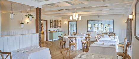 A restaurant with multiple tables and chairs, white tablecloths, hanging plants, and pendant lighting.