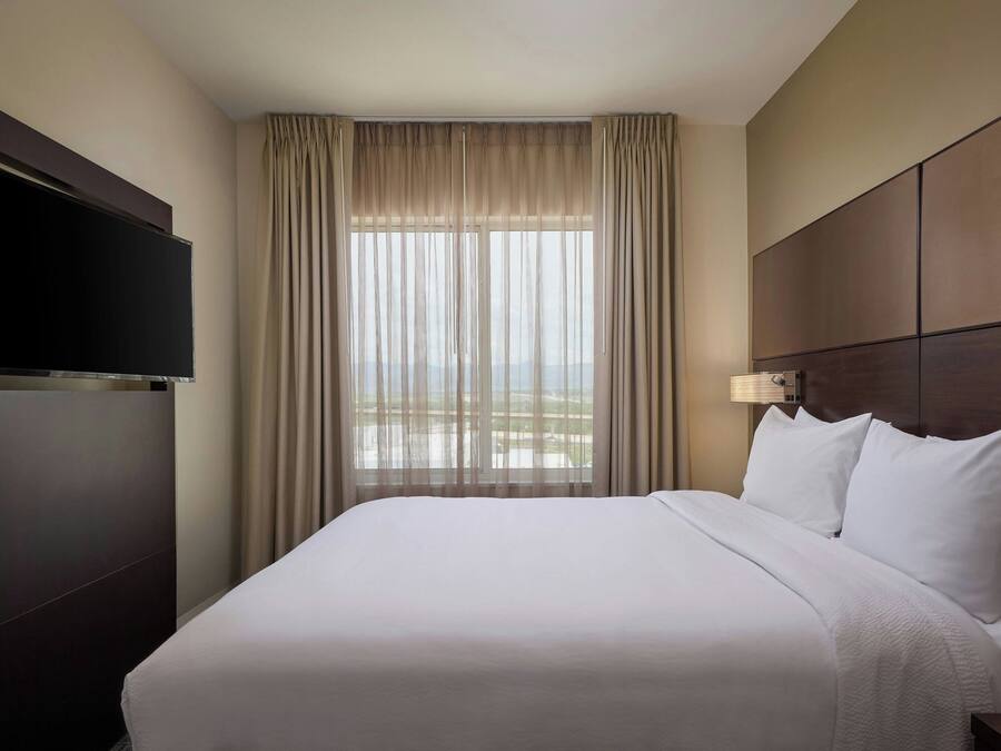 A hotel room with a large bed, a flat-screen TV, a window with curtains, and a nightstand with a lamp.
