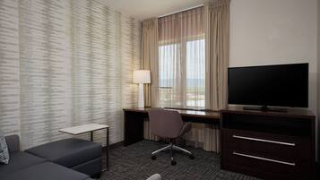 Quarto de hotel com sofá, escrivaninha com luminária, televisão e janela com cortinas.
