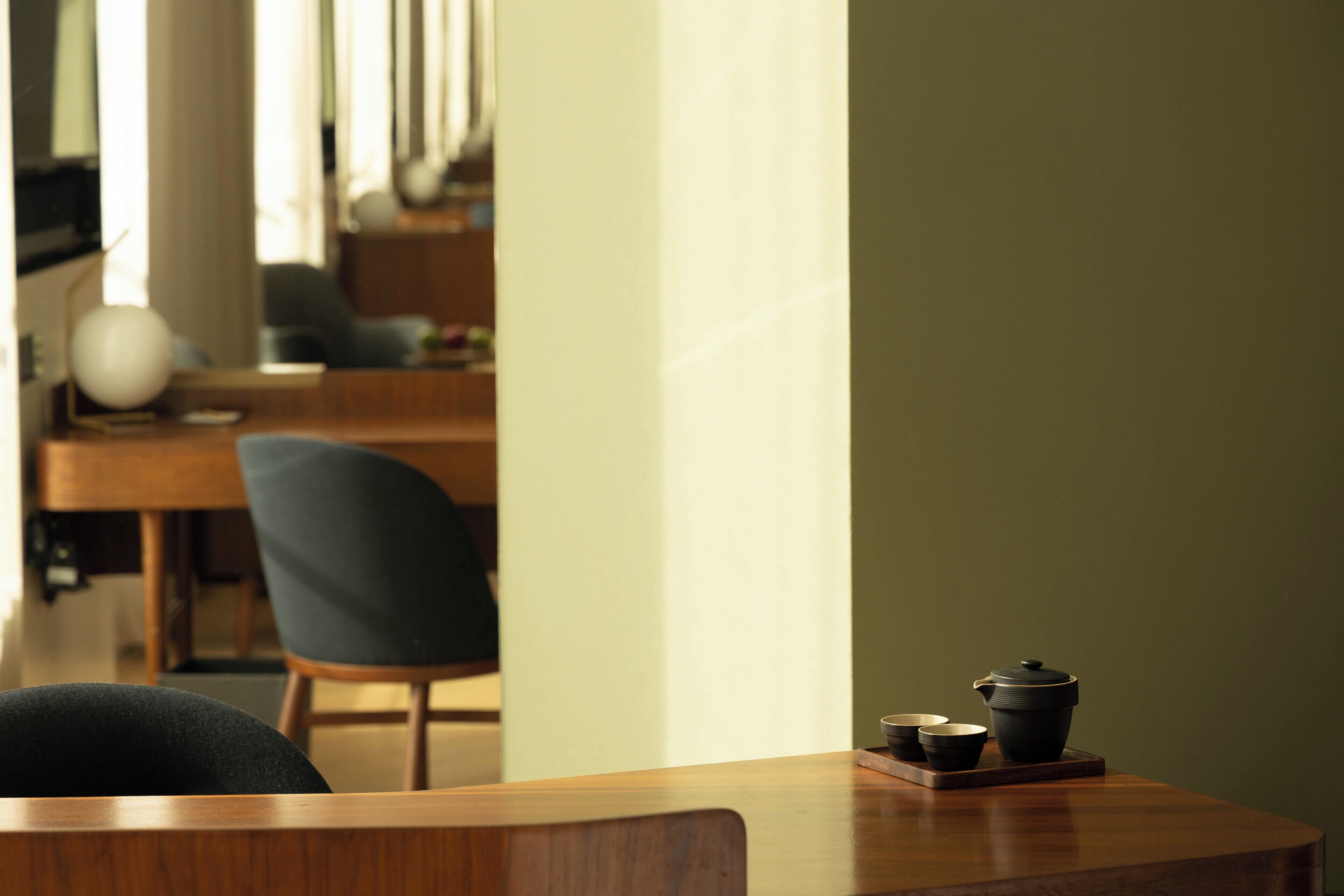 A wooden table with a teapot and cups, a chair, and a lamp in a room with a window and a chair in the background.